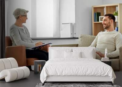 psychology, mental therapy and people concept - smiling senior woman psychologist with clipboard talking to young man patient at psychotherapy session Wall mural