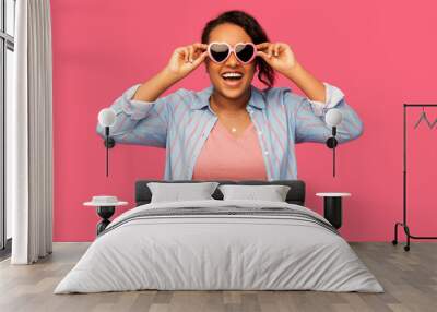 summer, valentine's day and eyewear concept - happy african american young woman in heart-shaped sunglasses over pink background Wall mural