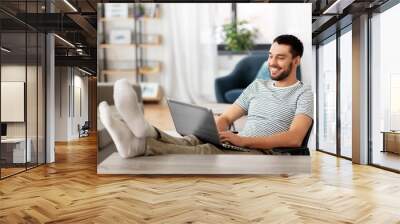 technology, remote job and lifestyle concept - happy smiling man with laptop computer resting feet on table at home office Wall mural