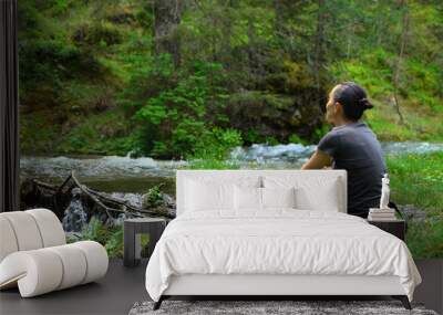 Young hiker woman sitting by a mountain stream, enjoys spending free time at nature Wall mural