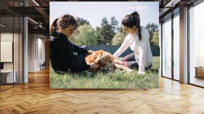 Low section view of girls and dog on ground. Portrait of dog lying on ground with grass and two girls petting him. Wall mural