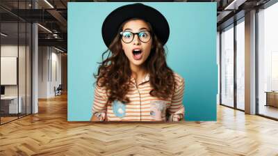 Young beautiful woman, fashion teen girl student wearing glasses and hat standing over blue background surprised and shocked with surprise expression, excited face Wall mural