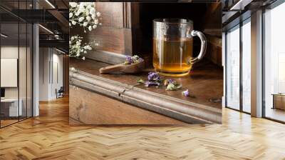 A transparent cup with herbal tea and a wooden spoon with dried herbs - thyme, green anise, ground ivy, pine bud, mallow flowers on an old wooden sideboard Wall mural