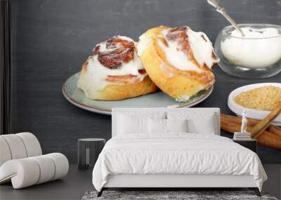 Two cinnabons buns lie on a green plate and next to cinnamon sticks and cane sugar in the right corner on a gray wooden background   Wall mural