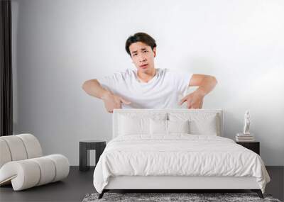 Doubtful and upset Young Asian Teenage boy wearing a white T-shirt standing over white background at home clueless and confused pointing down and looking to camera. Doubt thinking and Pensive concept Wall mural