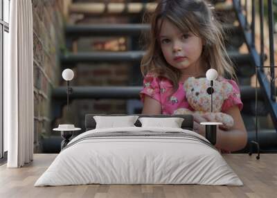 A young girl sits quietly on the stairs holding a teddy bear. Her serious expression captures a moment of reflection and innocence. Wall mural