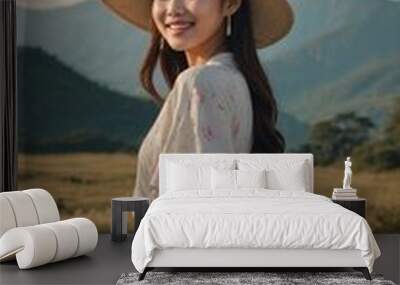 Young asian woman with long dark hair wearing wide-brimmed hat, standing in grassy field with mountains in background Wall mural