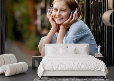 Pretty preteen child girl wearing headphones sitting near fence at city street and smiling. Cute female model with music earpods summer portrait Wall mural