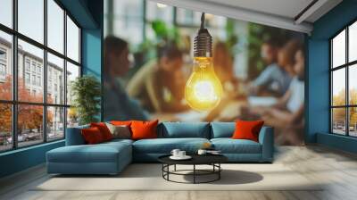  A light bulb hangs over the table in a group of people working together, representing the concept of innovation and creativity. The background is blurred, depicting an office interior. Wall mural