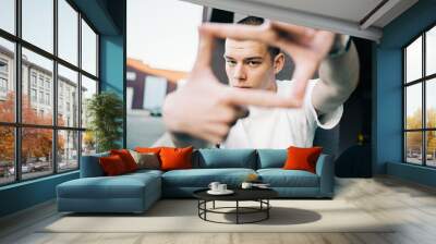 Handsome young man in early 20s looks into camera through a frame formed with his fingers. Millenial with short hair outdoors urban portrait. copy space for text. People concept Wall mural