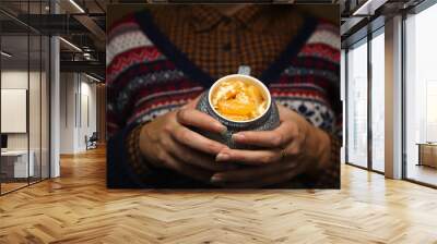 Closeup of female hands with a mug of creative beverage of mandarin orange lobule. Young hipster woman in Christmas sweater and shirt holding cup of tea or coffee. Cozy warm winter detail Wall mural