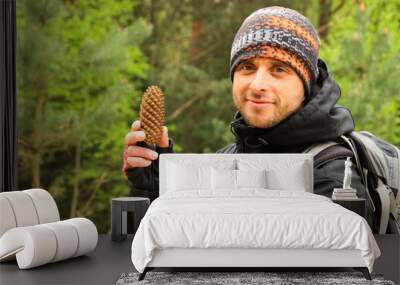 Young attractive male tourist with unshavenness in a sports jacket, backpack and buff in the autumn in the woods holds a pine cone in his hands and smiles. Wall mural