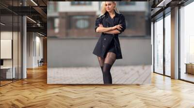 Beautiful blonde girl in a black coat with perfect legs in black pantyhose and shoes with high heels posing outdoor at the city square. Wall mural