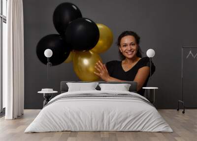 Cheerful African woman with beautiful smile poses with inflated black and gold air balloons against gray wall background with copy space. Concept of Black Friday and gold credit shopping card Wall mural