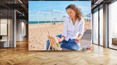 Happy young woman enjoying drawing on canvas in open air on the beach on warm sunny day Wall mural