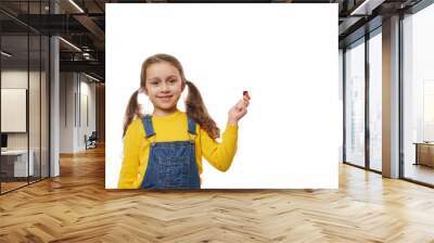 Isolated portrait on white background of happy child girl with two ponytails, wearing denim overalls and yellow pullover, cutely smiling looking camera, holding party horn, pipe, whistle in her hand Wall mural