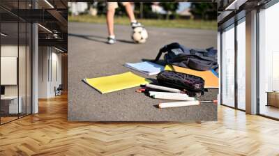 Scattered school supplies and workbooks falling out of an open backpack, lie on the asphalt of the schoolyard against the background of a boy's feet on a soccer ball. Wall mural