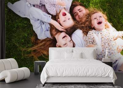 Happy cheerful family with two little cute daughters having fun and lying on the grass in the park. Sunny summer day. Parenthood, childhood, happy family concept. Outdoor Wall mural