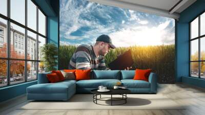 A young agronomist holds a folder in his hands on a green wheat field. A farmer makes notes on the background of agricultural land during sunset. Man in a cap with a folder of documents Wall mural