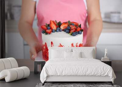 Pastry chef confectioner young caucasian woman in the kitchen. She holds out a berry-decorated with strawberries and blueberries cake on a plate. Front view. Сlose-up Wall mural