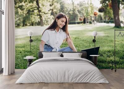 student girl with laptop outdoors. Smiling woman sitting on the grass with a computer, surfing the Internet or preparing for exams. Technology, education and remote work concept. Soft selective focus. Wall mural