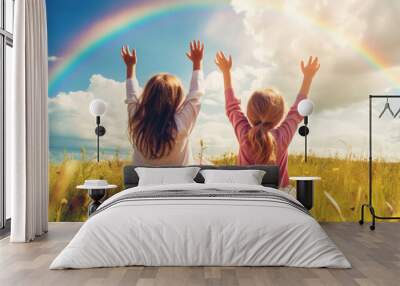 Two children with arms raised high, sitting in a meadow, reaching towards a vibrant rainbow. Wall mural