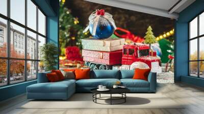 A red and white toy truck is going along a snow covered road against the background of festive lights and pines and carrying gifts. Concept of Christmas mood and preparation for the celebration Wall mural