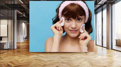 an attractive woman stands on a blue background in a pink soft rim with two cotton pads, I wipe my face with them, happily looking at the camera Wall mural