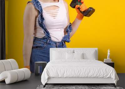 Builder girl in denim overalls and a black cap holds a drill and a screwdriver in her hands and poses on a yellow background Wall mural