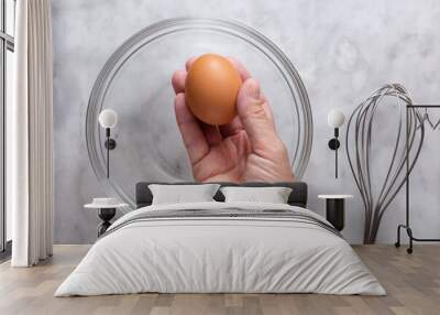 Top view of right woman hand holding one raw brown egg above glass bowl and whisk on marble surface Wall mural