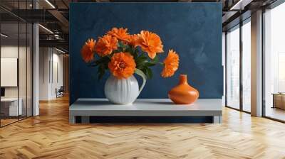 Minimalist decor featuring orange flowers in a white vase, an orange glass, and a blank card on a plate, set against a deep blue wall. Wall mural