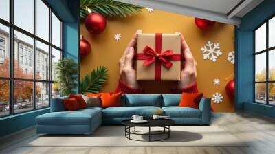 The hands of a man in a red sweater hold a green gift box with a red bow, against a background of small gifts, balloons and snowflakes. New Year's atmosphere with fir branches Wall mural