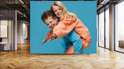 two cute, joyful children in multi-colored sweaters stand on a blue background, a boy rolls a happy girl on his back. The theme of children's friendship and relationships Wall mural