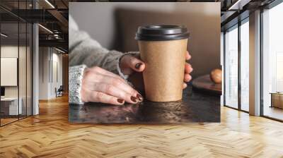 Women's hands in a warm sweater are holding a paper cup of aromatic coffee. The nails are coated with brown nail polish. Coffee manicure. Breakfast or a break in a cozy cafe Wall mural