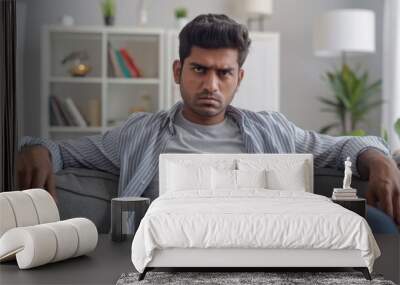 Young Indian man in casual attire, sitting on a sofa at home with a serious expression, gazing at the camera. Wall mural