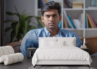 Young Indian man in casual attire, sitting on a sofa at home with a serious expression, gazing at the camera. Wall mural