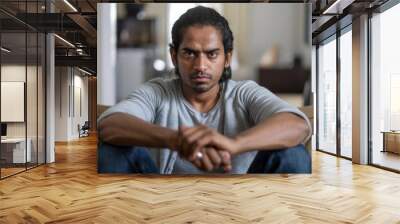 Young Indian man in casual attire, sitting on a sofa at home with a serious expression, gazing at the camera. Wall mural