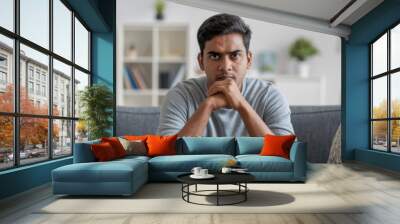 Young Indian man in casual attire, sitting on a sofa at home with a serious expression, gazing at the camera. Wall mural