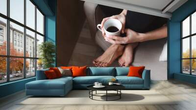 A young woman with a mobile phone is sitting in a comfortable chair by the window of the house and drinking tea. Home interior design. Freelancer Wall mural