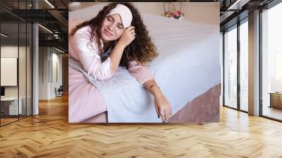 A beautiful curly-haired girl in a white sleep mask in pink silk pajamas lies on the bed, relaxed. A middle-aged woman is getting ready for bed in the morning, smiling. Wall mural