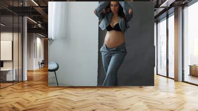 A beautiful pregnant brunette woman in a cropped gray stylish suit on a gray paper background in the studio. A pretty girl with a bare stomach in a fashionable suit is sitting barefoot on a gray backg Wall mural