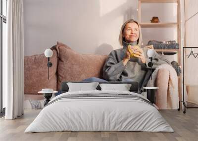 Happy dreamy middle aged woman sitting on sofa in living room with cup of tea or coffee Wall mural