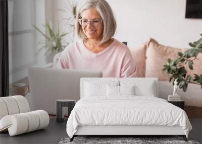 Happy middle age senior woman sitting at the table at home working using computer laptop Wall mural