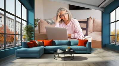 Sad middle-aged woman sit at table in living room using laptop Wall mural