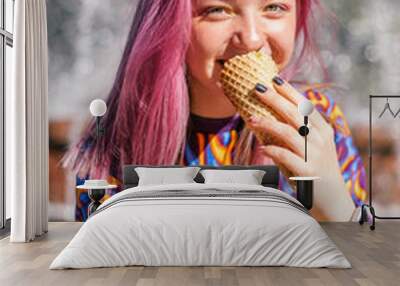 Happy pink-haired teenage hipster girl in a colorful bright T-shirt is eating ice cream on a summer day, with a city fountain in the background.Summer concept.Generation Z style. Wall mural