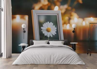 A white picture frame with a single white daisy flower inside rests on a table with a row of candles burning in the background. Wall mural