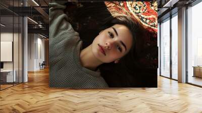 A young woman with long brown hair lies on a patterned rug, gazing directly at the camera. Her sweater is grey and the light is soft, highlighting her features. Wall mural