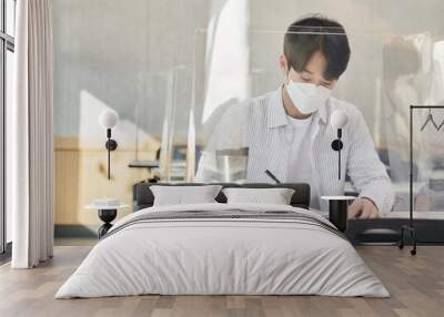 A young male Asian college student is learning or listening to a lesson while her face is outside a protective shield on the desk in a university classroom in Korea during the pandemic. Wall mural