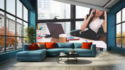 business woman talking on the mobile phone and smiling while sitting at her working place in office and looking at laptop Wall mural