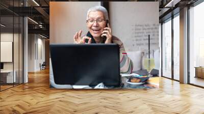 Happy senior old woman sitting in a coffee shop and doing some work on her laptop or browsing the internet Wall mural